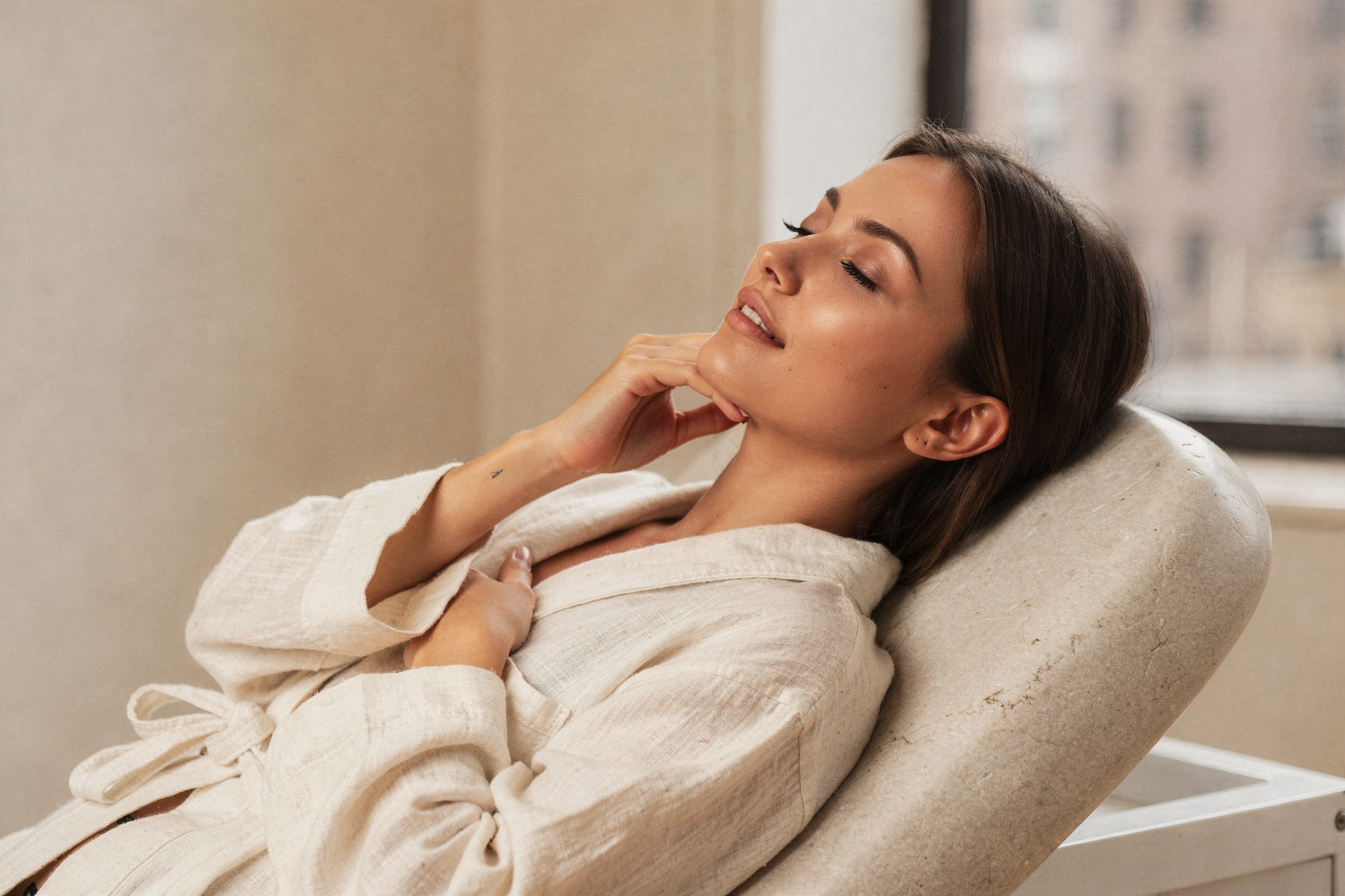 Woman relaxing on a beige chair in a bright room with city views, touching her face
